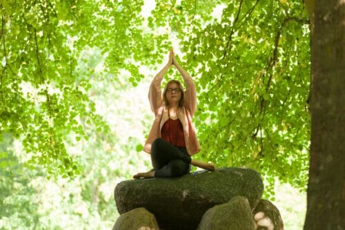 Cathrine sidder på en stor sten i en grøn bøgeskov og laver meditativ yoga