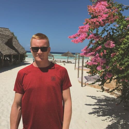 Tommy står på stranden ved busk med lyserøde blomster på yogarejse til Zanzibar