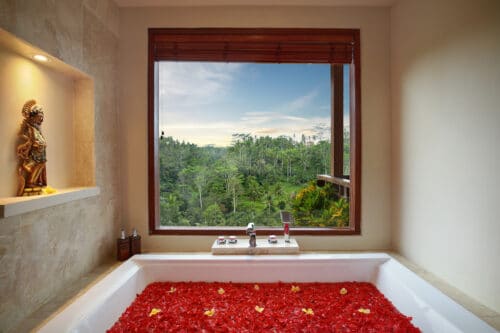 Badeværelse med badekar der er fyldt med blomsterblade og stort vindue med udsigt til junglen på yoguddannelse på Bali